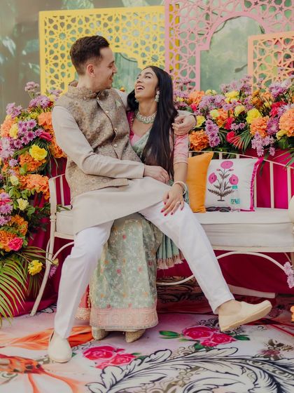 A playful and candid moment from a pre-wedding ceremony. The couple shares a laugh while seated on a bench against a vibrant floral backdrop, showcasing their fun-loving personalities.