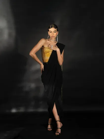 A full-length shot of the black and gold draped dress from the 'Gleam Collection'. The model's pose is directed to showcase the outfit's structure and modern aesthetic.