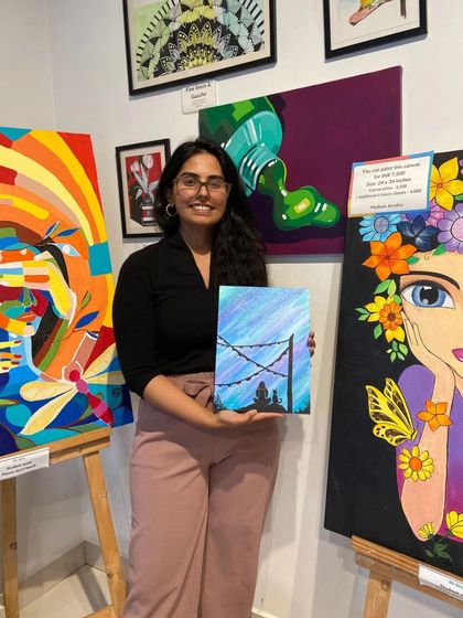 A participant with her finished canvas painting, surrounded by other beautiful artworks in the studio.