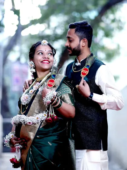 A happy couple shares a candid moment during their outdoor baby shower photoshoot. The mom-to-be is adorned in traditional floral jewelry, and they both wear playful 'Aai' (Mom) and 'Baba' (Dad) badges.