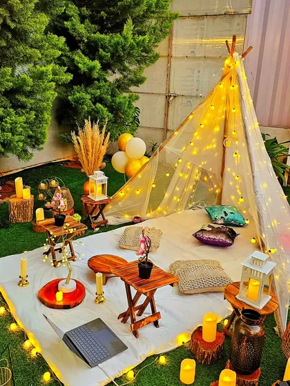 A close-up of a beautifully styled boho picnic at night. The setup includes a teepee tent wrapped in fairy lights, low wooden tables, plush cushions, and an array of candles and lanterns for a warm and inviting atmosphere.