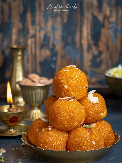 A pyramid of Motichoor Ladoos, glowing in the light of a diya. The dark, rustic background makes the orange hue of the ladoos stand out, creating a classic festive image.