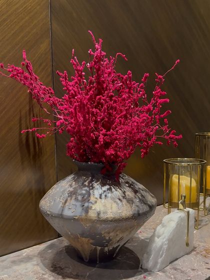 A bold and modern decorative piece. A rustic, textured vase holds a vibrant arrangement of dried pink foliage, complemented by minimalist gold and marble candle holders.