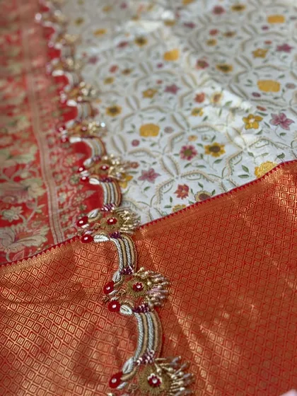 An ornate border design with intricate zardosi, beadwork, and red accents on a white floral brocade, paired with a red silk saree.