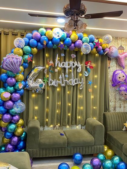 An underwater adventure with a Little Mermaid theme for a fourth birthday. This living room decoration includes a balloon arch in ocean colors, mermaid tail and seashell foil balloons, and fairy lights behind a green curtain to create a shimmering sea effect.