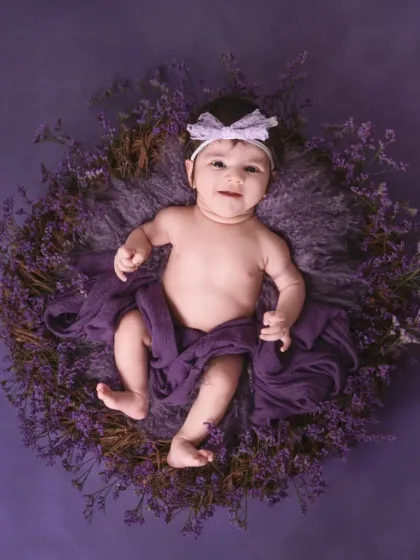 This adorable baby girl is nestled in a wreath of purple flowers against a solid purple backdrop. Her sweet smile and direct gaze make this a captivating infant portrait.