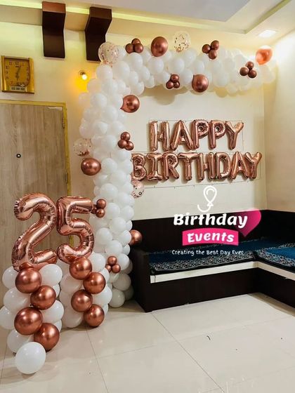 A clean and classic 25th birthday decoration for a living room, featuring rose gold foil and number balloons with a white balloon arch.