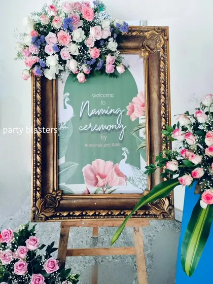 This custom welcome sign for a naming ceremony features a gentle peacock and floral design. The ornate gold frame is adorned with a beautiful arrangement of fresh roses and baby's breath, setting an elegant tone for the event.