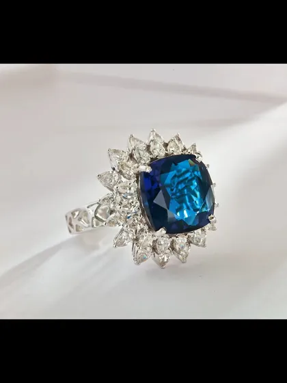 A close-up of a magnificent cushion-cut blue sapphire ring, framed by a brilliant halo of marquise and round diamonds. The studio lighting is carefully controlled to highlight the deep, saturated color of the sapphire and the fire of the surrounding diamonds, making it an ideal shot for a luxury brand's homepage or product detail page.