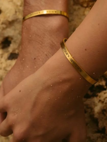 An intimate shot of a couple holding hands, their matching gold bracelets with custom engravings catching the light. The permanent engraving ensures your story lasts a lifetime.