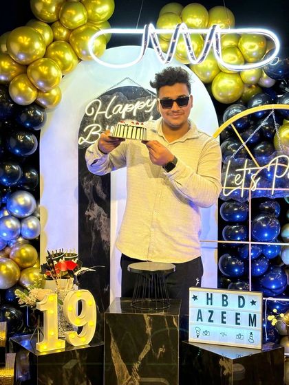 A cool pose with the birthday cake. The black and gold theme with "Let's Party" and "Happy Birthday" neon signs provides a vibrant and fun backdrop, perfect for a 19th birthday celebration.