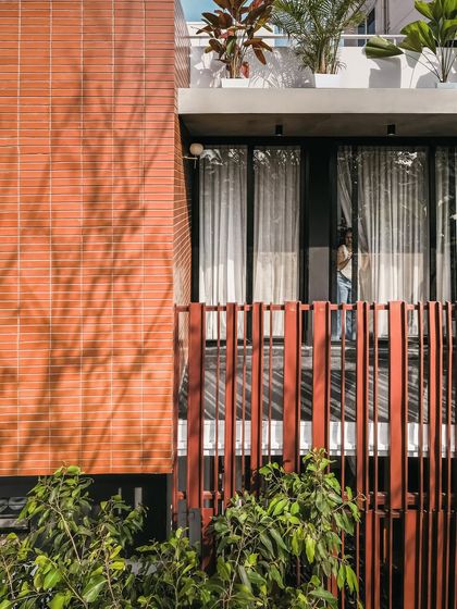 The combination of terracotta brick, dark window frames, and the rhythmic pattern of the vertical screen creates a rich textural palette on the 'Ittige' facade. The play of shadows from the nearby trees adds another layer of dynamic beauty.