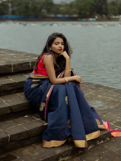 A pensive portrait by the water. The model sits on stone steps, her thoughtful expression and the calm water behind her creating a serene and contemplative mood for this saree photoshoot.