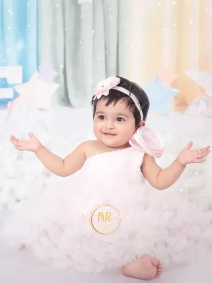 A happy baby girl with her arms outstretched, sitting in a whimsical cloud and rainbow setup for her first birthday photos.