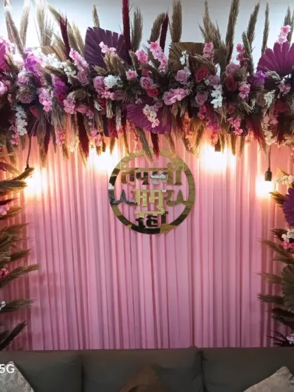 A dense floral arch with pink and purple flowers mixed with pampas grass. This backdrop for a Jain ceremony features a golden sign and is set against a pink curtain.