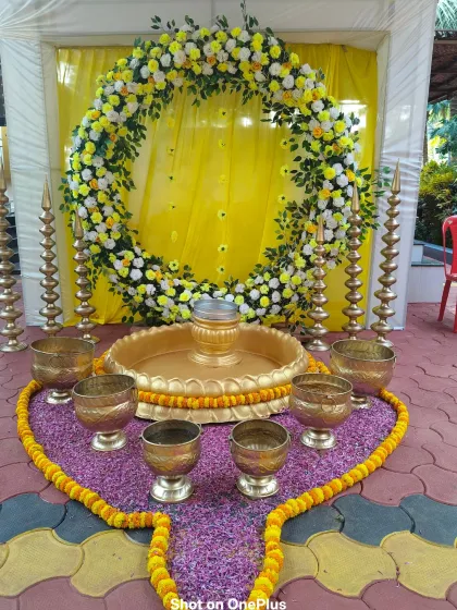 An outdoor Haldi setup in Kundapura. The ritual area is decorated with a floral carpet of purple petals, marigold borders, and a circular floral arch in yellow and white.