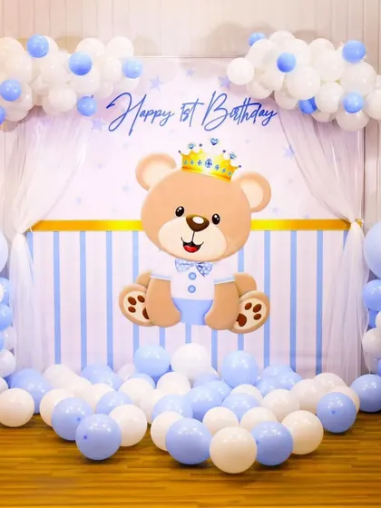 A sweet teddy bear theme for a first birthday, perfect for a baby boy. The backdrop features a cute crowned bear, with balloon columns and clouds in a soft blue and white palette.