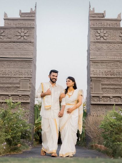 A couple dressed in elegant white and gold traditional South Indian attire, standing before a monumental stone structure, creating a powerful and majestic image.