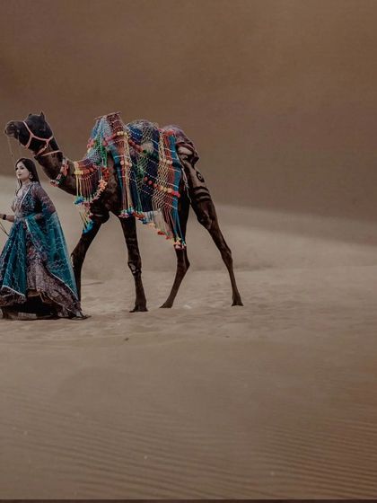 A beautiful portrait of the bride-to-be in a traditional blue outfit, walking alongside a camel in the desert. The flowing fabric and golden sand create a stunning visual.