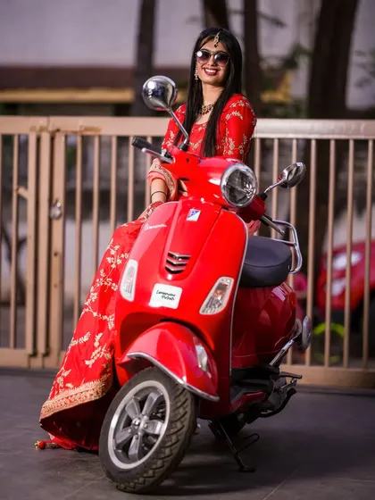 A cool bride in a red lehenga and sunglasses, posing confidently on a vibrant red Vespa. A perfect mix of tradition and modern swag.