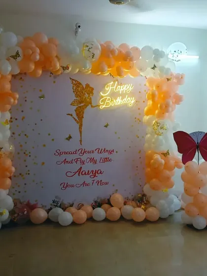 A fairy and butterfly theme for a first birthday. This setup includes a personalized backdrop, a peach and white balloon arch, and a "Happy Birthday" neon sign.