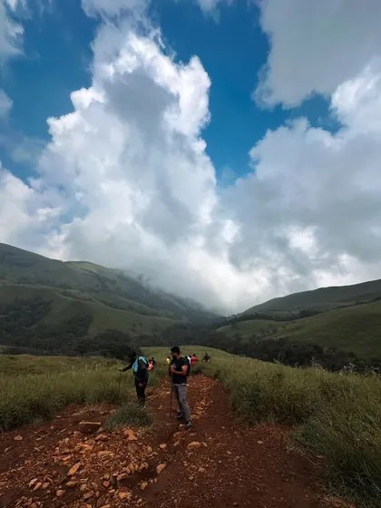 Hitting the trails at Netravathi peak. The path winds through vast green meadows under a dramatic cloudy sky.
