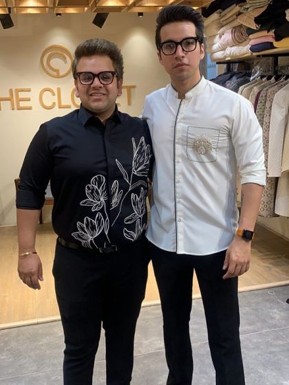 My brother Nikhil and I, the founders of The Closet. We are wearing two of our custom shirt designs, a black floral embroidered shirt and a white asymmetrical zip shirt, showcasing our unique approach to smart casual wear.