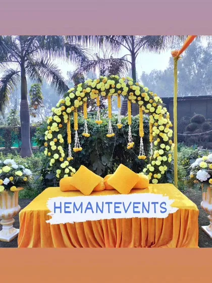A bright and sunny outdoor Haldi seating setup. The yellow divan is complemented by a matching floral arch and classic white urns filled with flowers, creating a cheerful atmosphere.