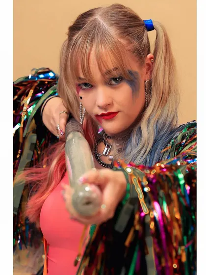 A close-up of the Harley Quinn cosplay, showing the character's signature pigtails, makeup, and a prop baseball bat pointed toward the camera.