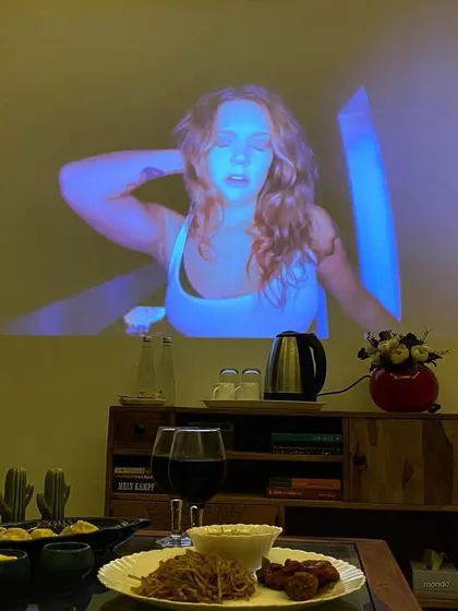 A dinner table set for two, with food and wine, while a movie provides the evening's entertainment on the wall.