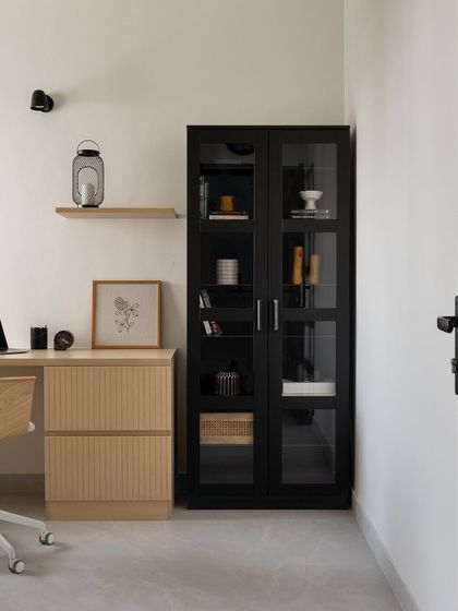A minimalist study area with a light wood desk, fluted drawers, and a sleek black glass-front cabinet for storage. The simple floating shelf and wall sconce keep the space uncluttered and focused.