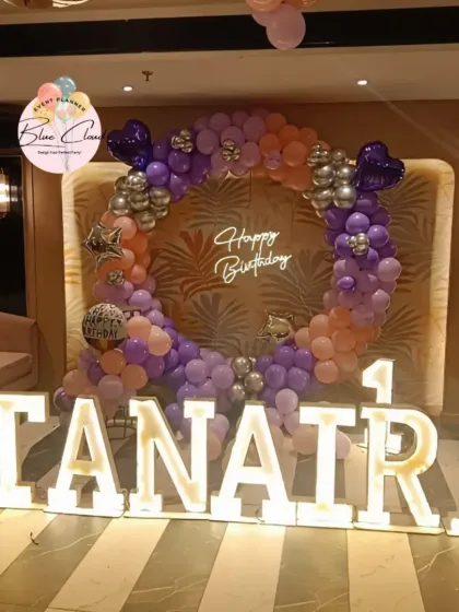 A large-scale first birthday decoration with marquee letters and a balloon ring in a party hall.