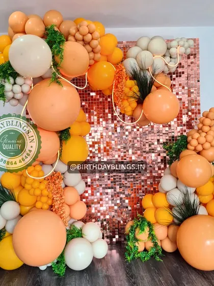 An impressive balloon installation with a rose gold shimmer wall, using a vibrant orange and white color scheme with faux greenery.
