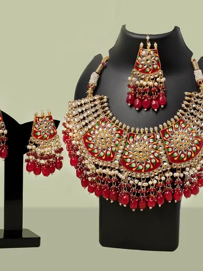 A professional catalog shot of a traditional red and gold bridal jewelry set, including a necklace and earrings, displayed on a bust for a clear view.