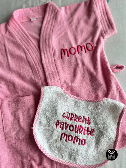 This thoughtful gift features a personalized pink bathrobe for a baby girl and a matching bib that says 'Current Favourite Momo'.