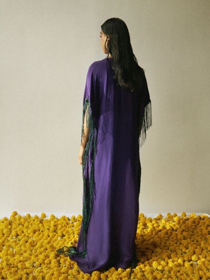 A quiet, contemplative moment from an editorial shoot. The styling focuses on the silhouette of a purple kaftan with green fringe, set against a bed of yellow marigolds, creating a rich color story with a minimalist composition.