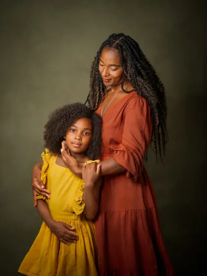 A mother's gentle touch and loving gaze toward her daughter. This portrait captures the tender, protective bond between them. The rich colors of their dresses against the painterly green background create a warm and vibrant image of family love.