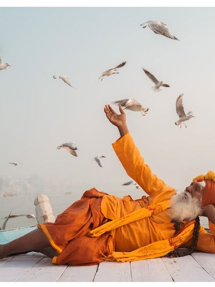 A sadhu reclines on a boat, one hand outstretched towards the flocking seagulls. This image captures a moment of effortless grace and connection with nature on the Ganga.