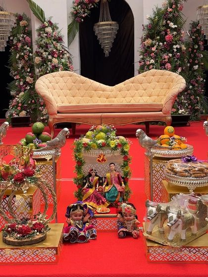 A modern and elegant baby shower setup with a rich red carpet and beautiful floral trees. The plate decorations are placed on stylish golden stands, and the scene includes charming dolls and props.