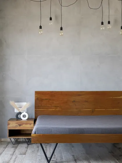 This bedroom combines an industrial aesthetic with rustic warmth. A custom wooden bed with metal legs is set against a concrete-finish wall, with exposed bulb lighting adding to the raw, edgy feel.