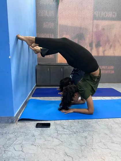 This student is working on her Scorpion pose (Vrischikasana) against the wall. This is an advanced inversion that combines a forearm stand with a deep backbend, requiring both strength and flexibility.