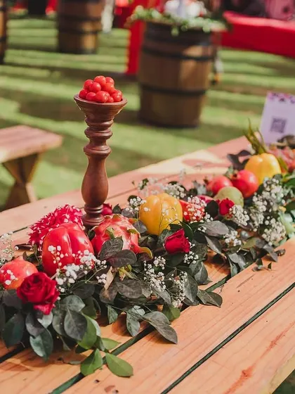 A rustic picnic table transformed with a stunning runner of fresh vegetables and flowers for a 'La Tomatina' themed Haldi. This centerpiece features red and yellow bell peppers, pomegranates, and roses, showcasing a creative and sustainable approach to event decor.