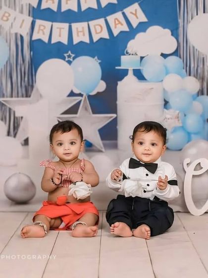 A first birthday celebration for twins. The blue and white 'Happy Birthday' backdrop with star and cloud decorations creates a festive and dreamy setting for this milestone sibling shoot.