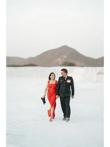 A wide shot of the couple walking hand-in-hand in a surreal white landscape, the groom in his uniform and the bride in a striking red dress.