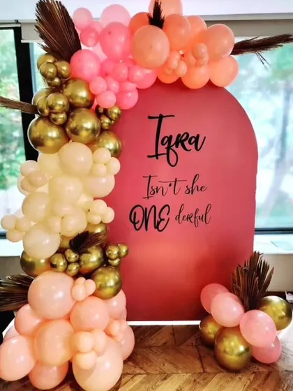 A modern and stylish first birthday backdrop. The simple arched board with custom text is decorated with a beautiful arrangement of peach, cream, and gold balloons.