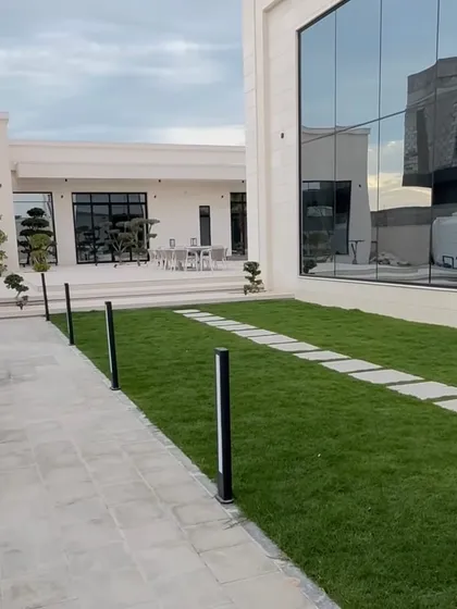 This view showcases the integration of hardscape and softscape elements. A neat lawn is bordered by a stone pathway with modern lighting, leading to a large glass wall that reflects the beautiful greenery.