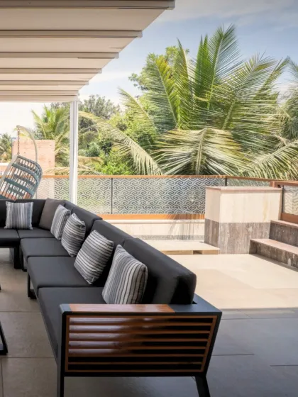 An outdoor lounge area designed for relaxation, featuring a comfortable sectional sofa and a hanging swing chair. The space is sheltered yet open, offering views of the surrounding palm trees and greenery.