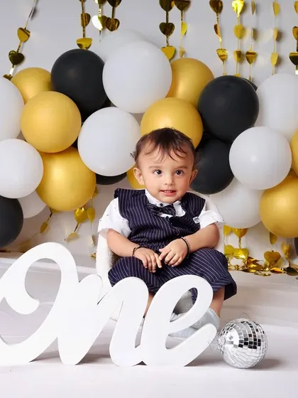 A chic and modern first birthday theme featuring a black, white, and gold balloon wall. The little gentleman in his vest looks ready to celebrate his big day in style.
