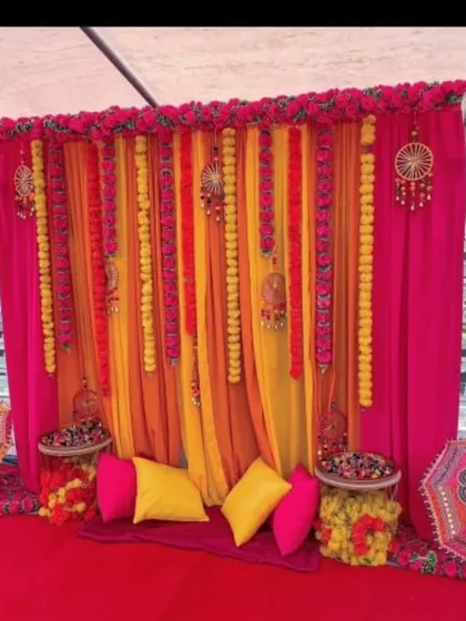 A cozy and colorful Mehandi seating corner created inside a tent. The backdrop of pink and yellow drapes is decorated with marigold garlands and traditional hangings, perfect for intimate gatherings.