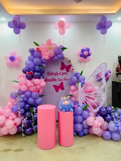 A lovely butterfly-themed setup in shades of purple and pink. This decor features a balloon arch, a large butterfly wing prop, and pedestals for the cake, perfect for a third birthday.